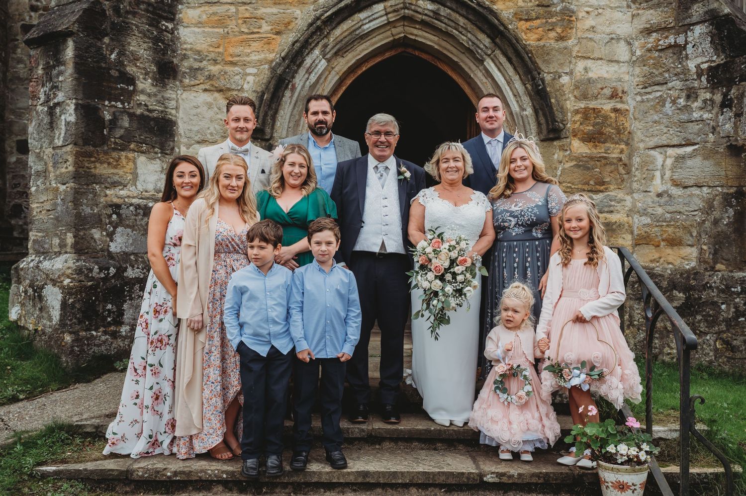 A wedding party poses together in front of a historic stone church archway entrance with floral decorations.