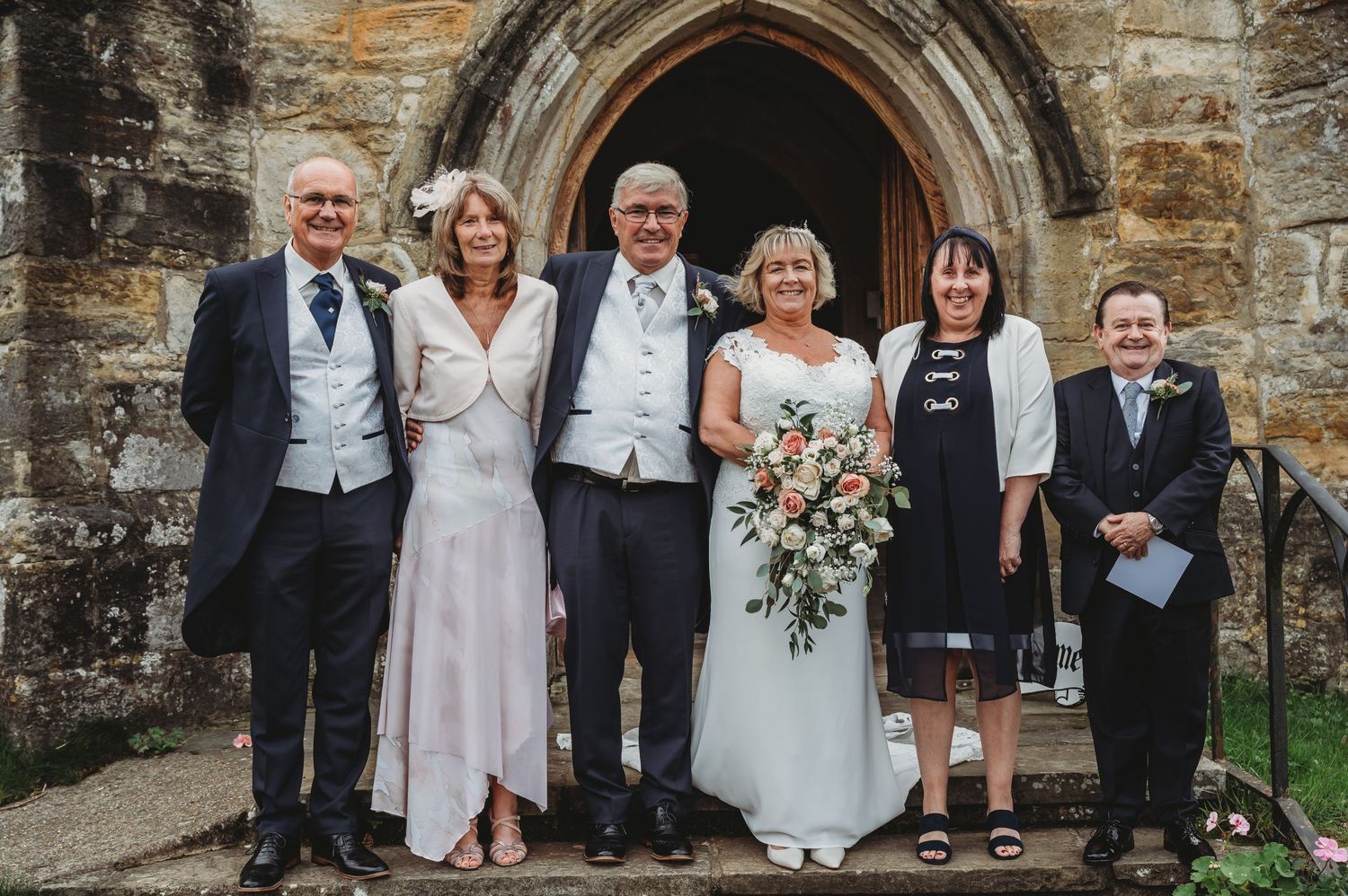Group in formal attire stands together smiling outside traditional stone church entrance with decorative archway.