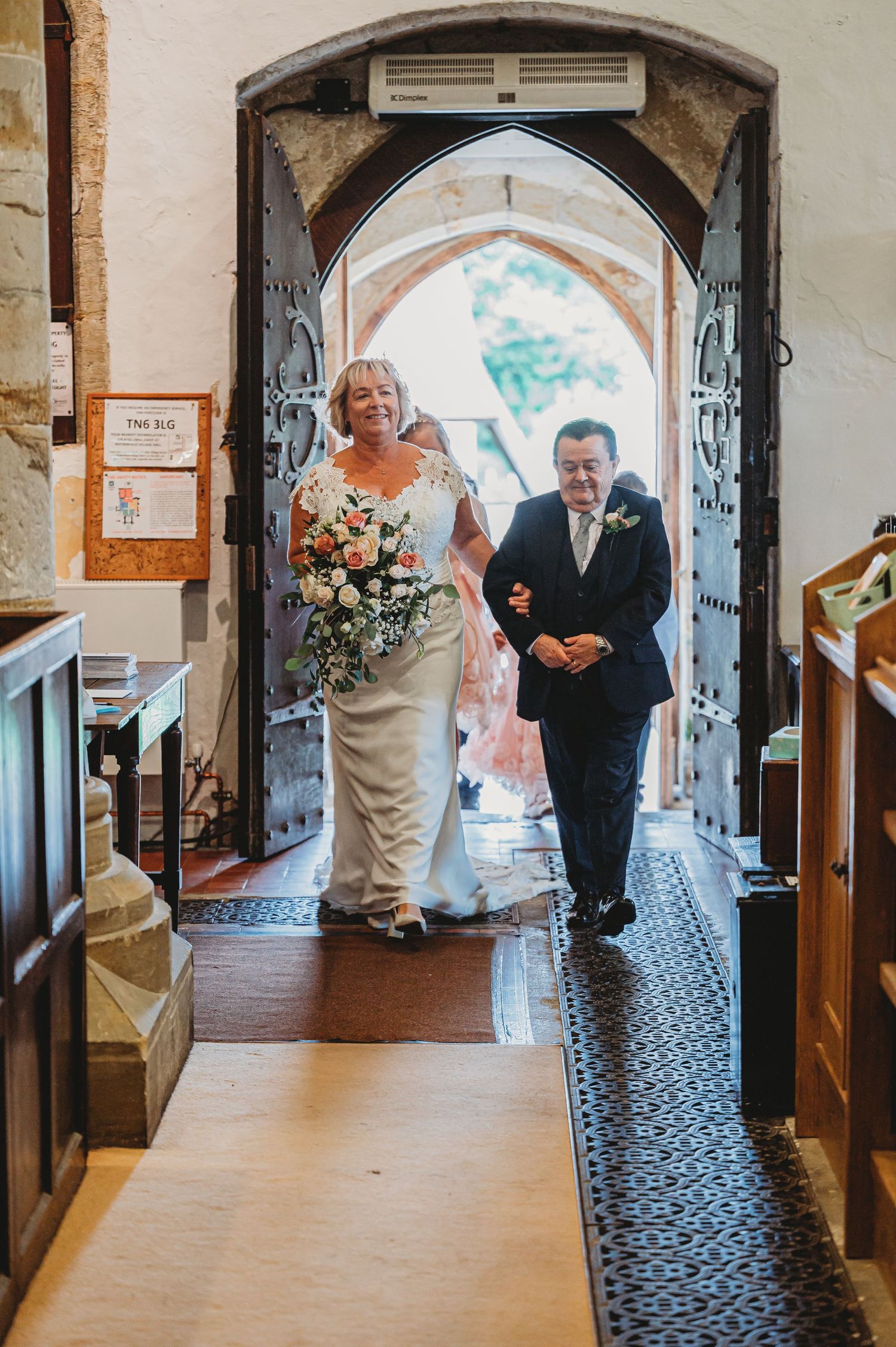 A couple walks down a church aisle together under an arched doorway with decorative details and carpeted flooring.