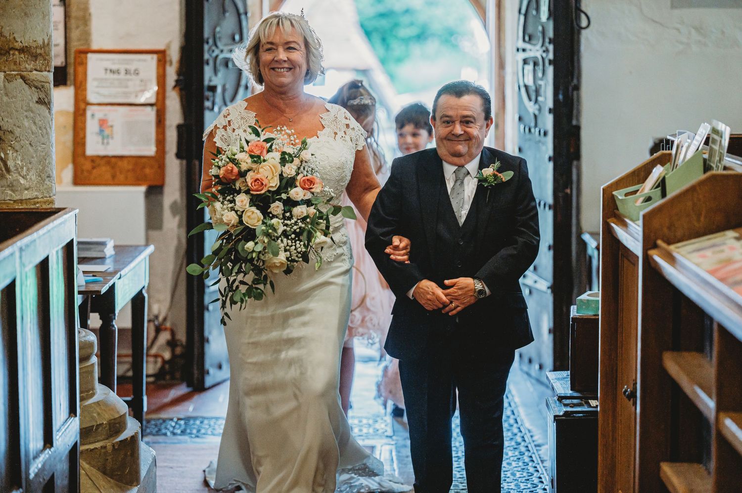 Wedding ceremony entrance procession inside ornate church with bright natural lighting and floral bouquet.