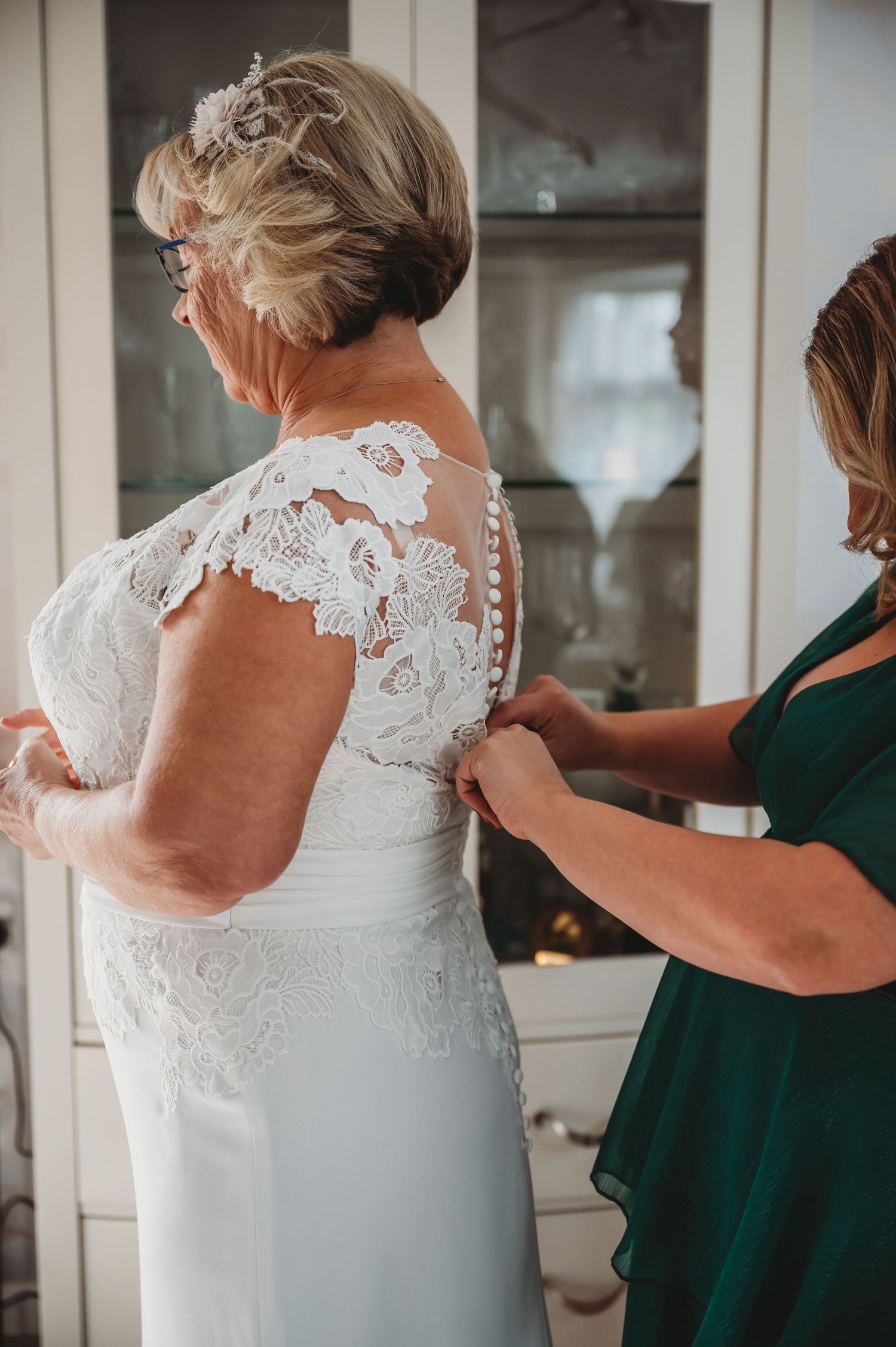 Someone in emerald green helps button up the delicate lace back of an elegant white wedding dress.