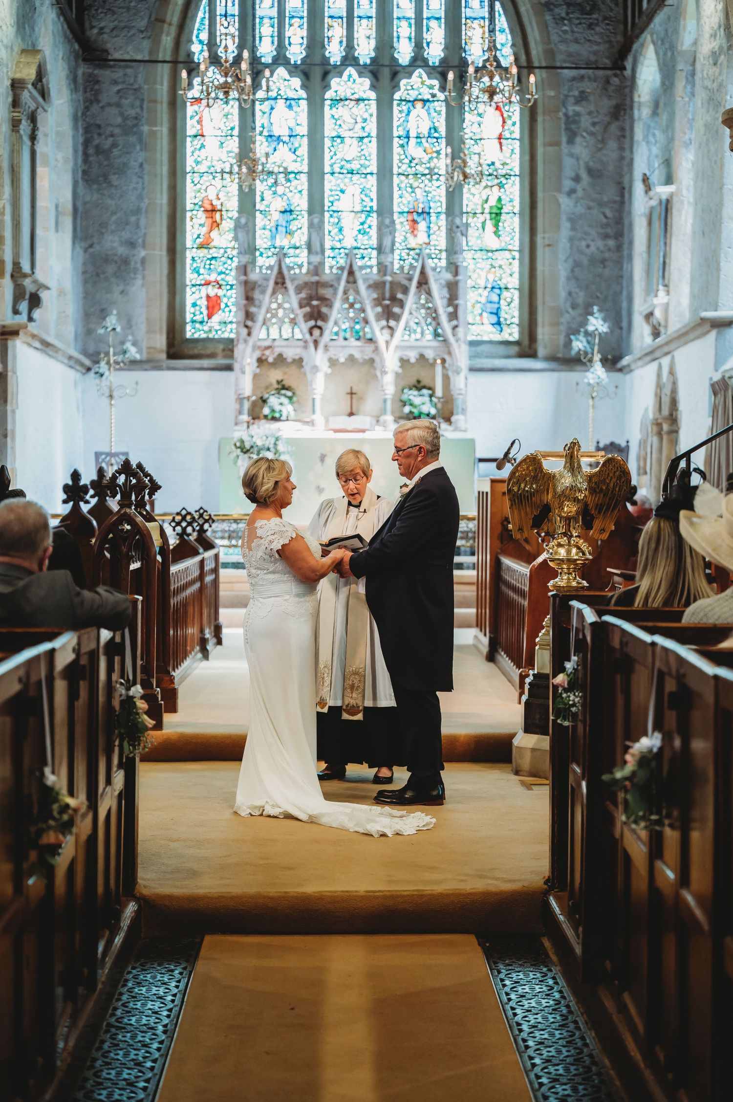 A wedding ceremony takes place in a historic church with stained glass windows and traditional wooden pews.