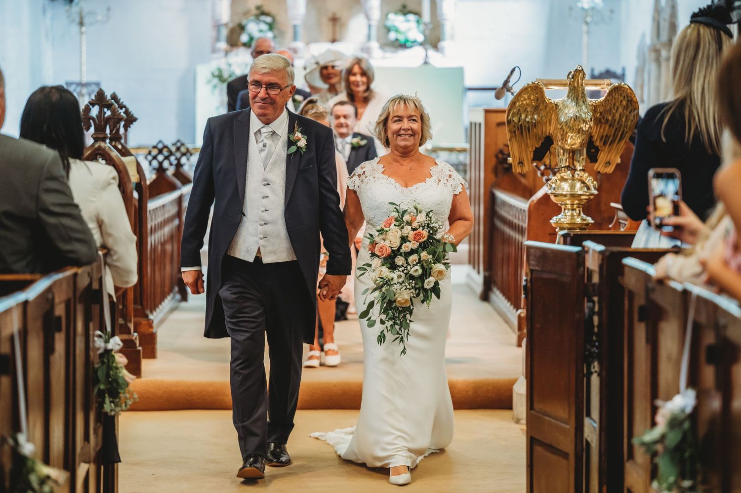 A bride walks down the church aisle with her escort at a wedding ceremony while guests capture photos.