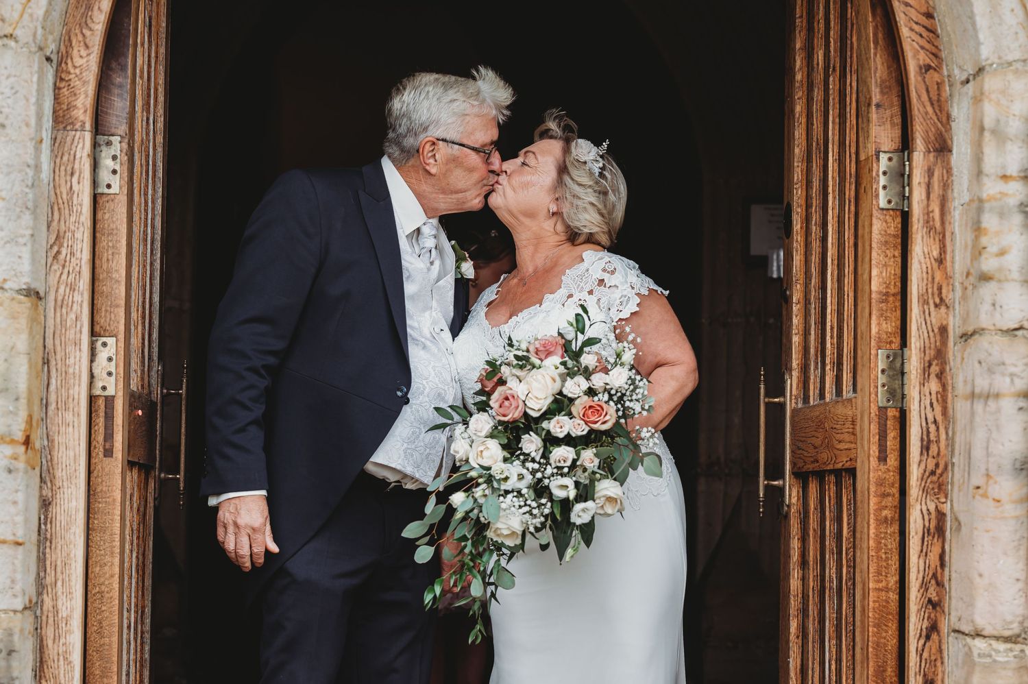 A happy wedding couple sharing a tender moment in a church doorway while holding a pink and white floral bouquet.