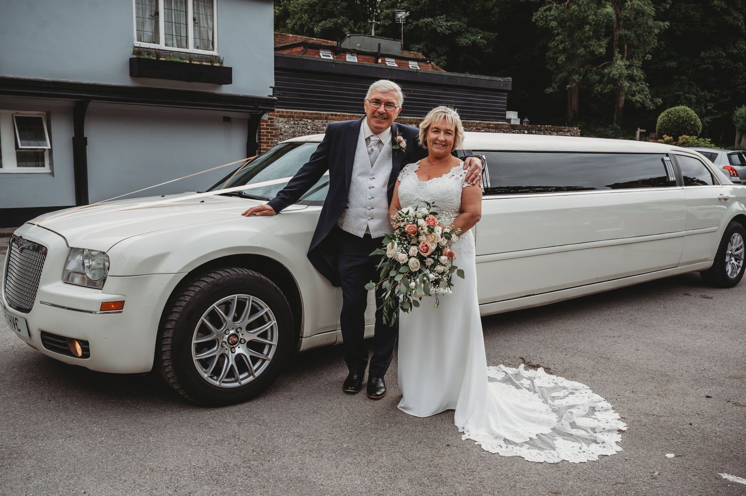 A white Chrysler stretch limousine parked outside with two people in formal attire posing beside it.