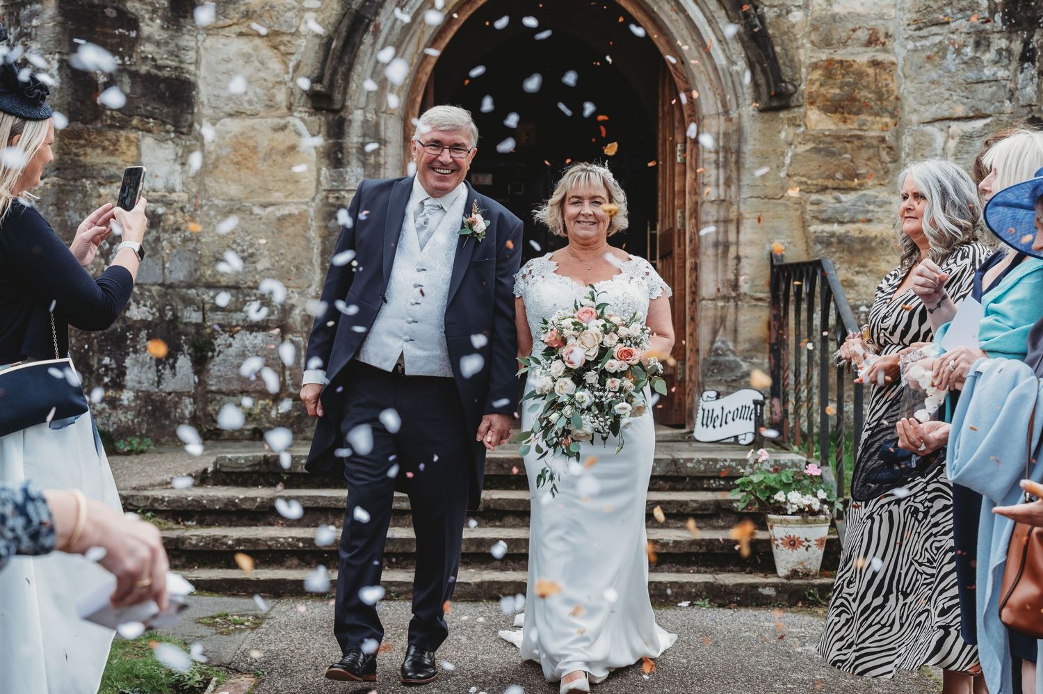 A wedding couple exits a stone church while guests throw confetti during a joyous ceremony celebration.
