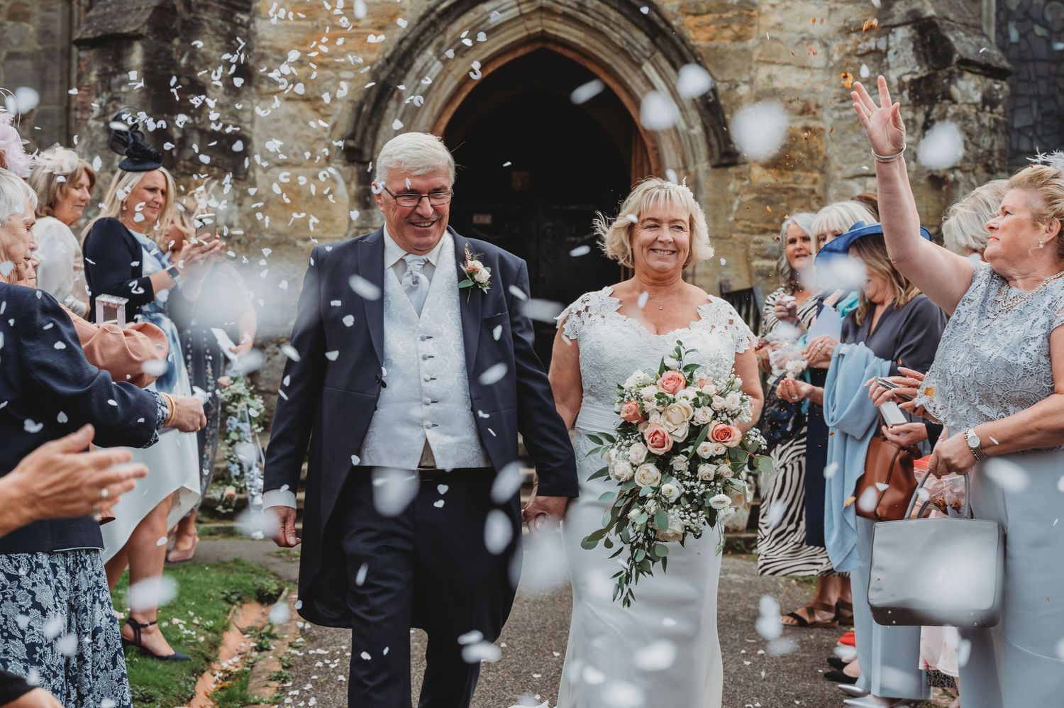 Wedding guests shower the newlywed couple with confetti as they exit a stone church after their ceremony.