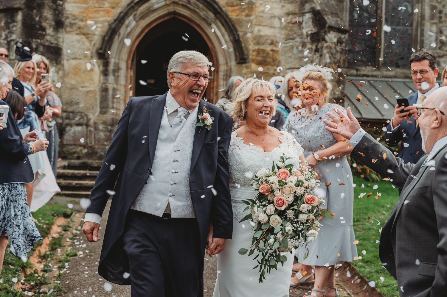 Joyful couple exits church while guests throw flower petals during wedding celebration.