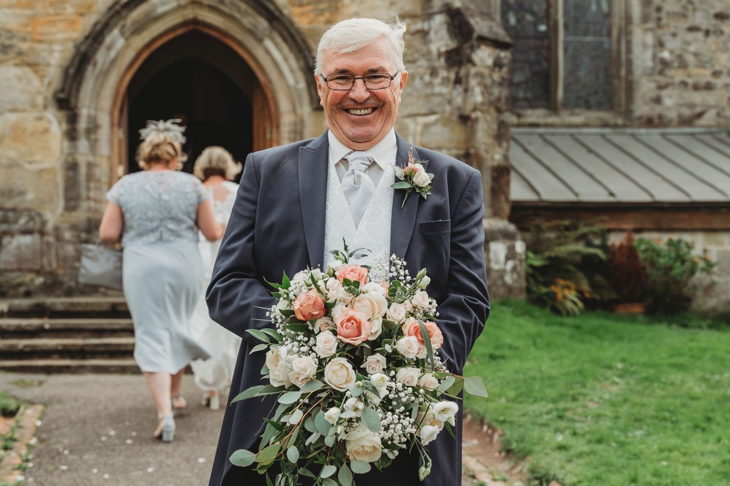 A person in a dark suit holds a beautiful wedding bouquet with pink and white flowers outside a stone church.