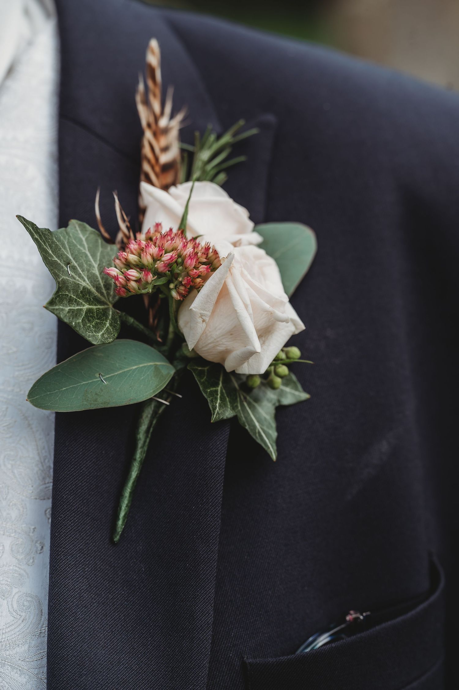 A delicate white rose boutonniere with feather accent and pink berries adorns a black suit lapel.