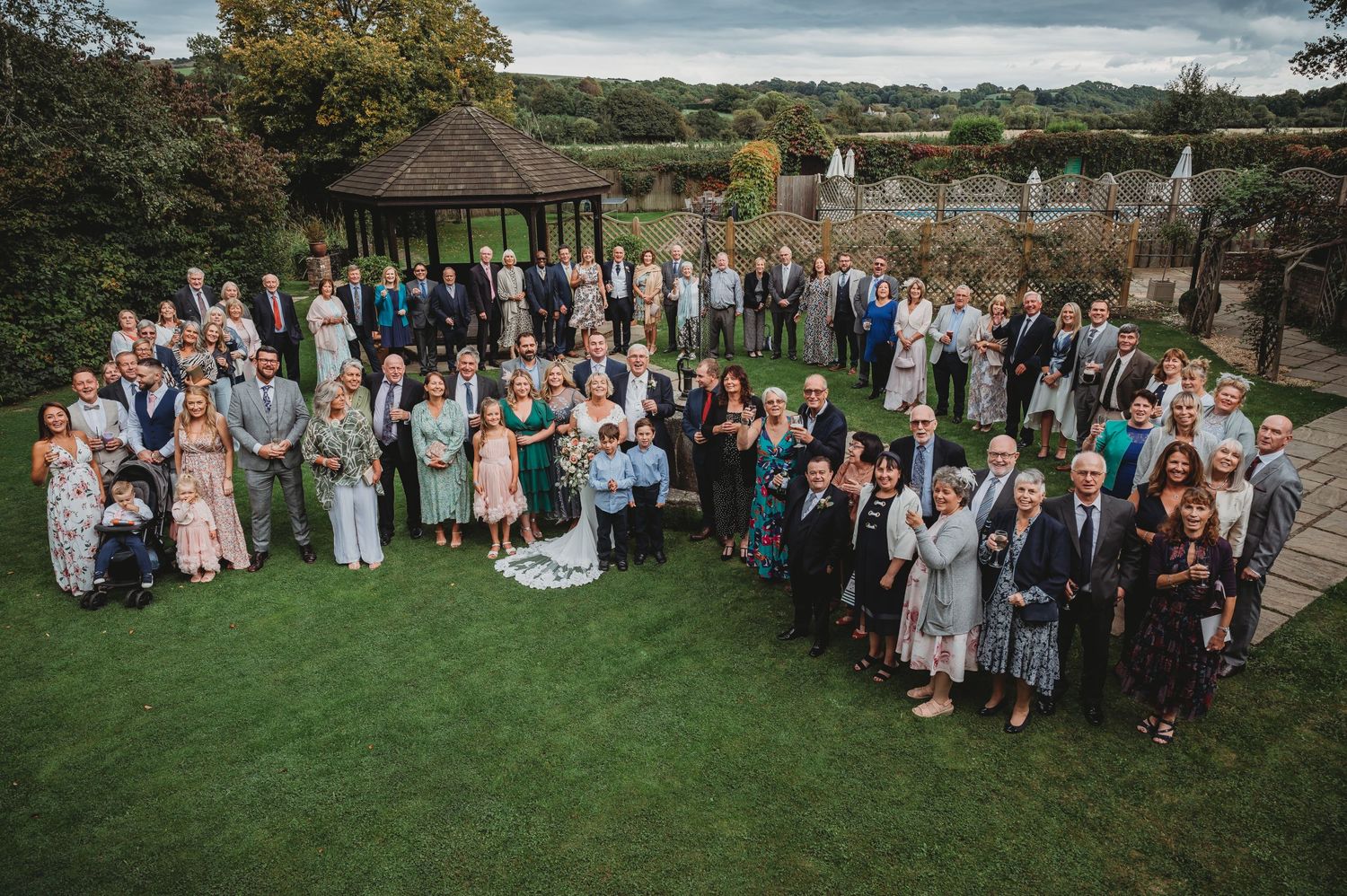 A large group of wedding guests gathered on a green lawn with a gazebo and scenic countryside view in the background.