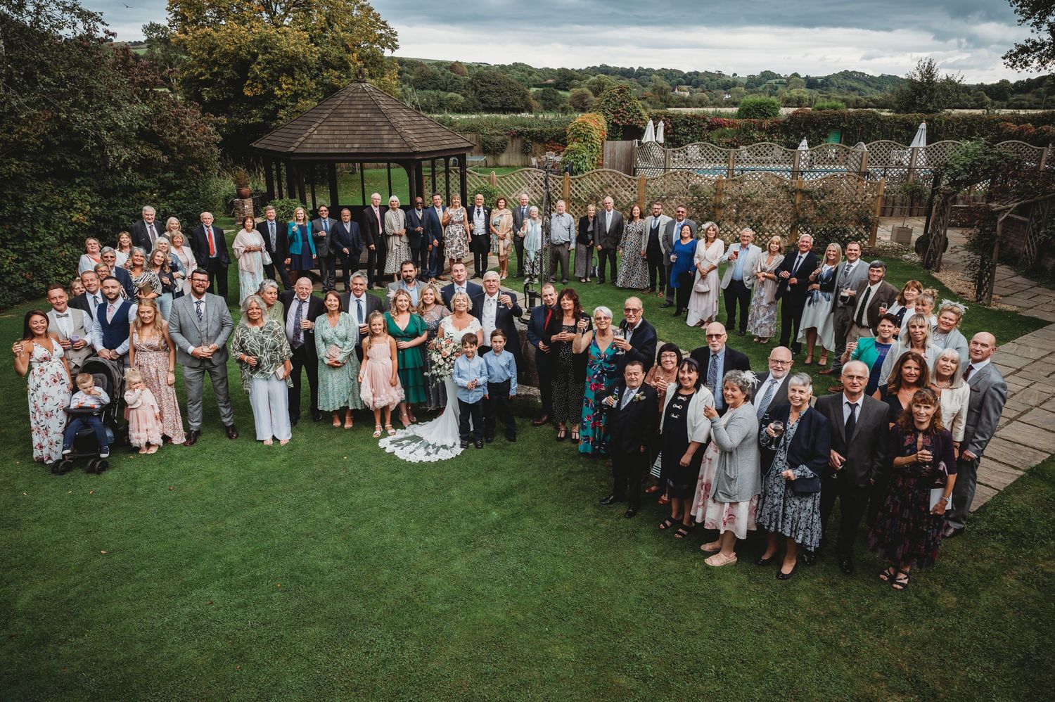 Large group of wedding guests gathered on a green lawn in front of a gazebo during an outdoor reception.