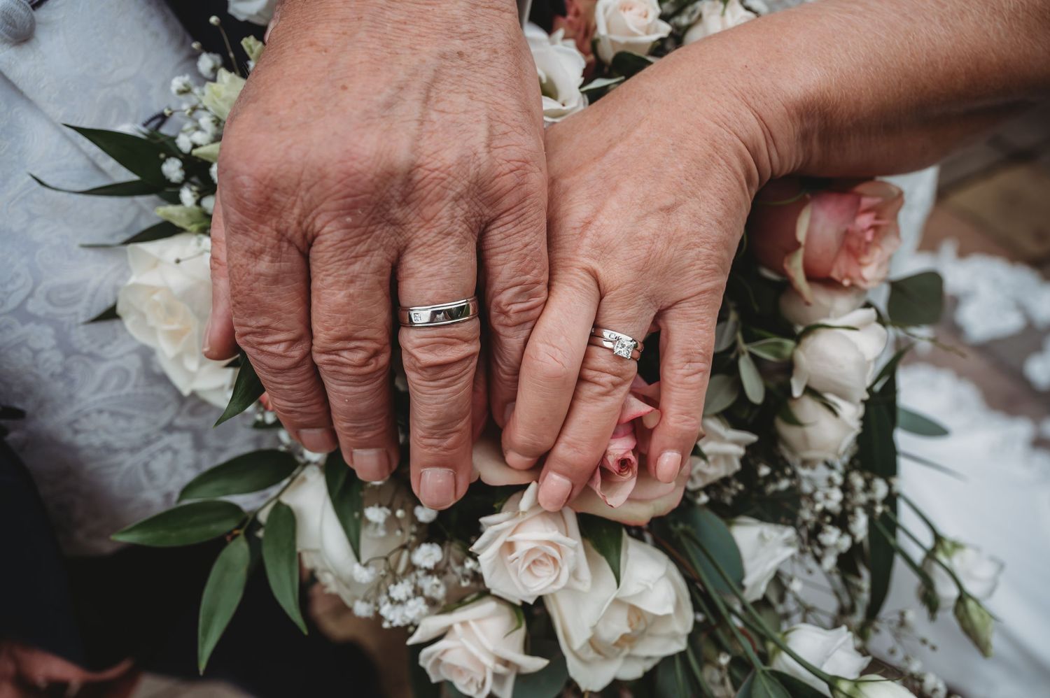 Wedding rings and manicured hands shown against a romantic bouquet of white roses and greenery.