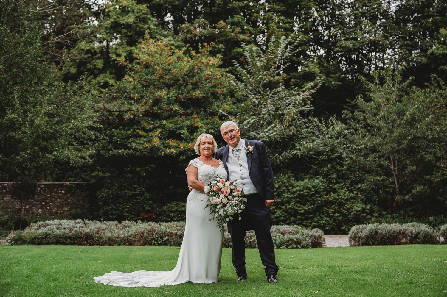 A wedding couple poses together on a manicured lawn with lush greenery and trees in the background.