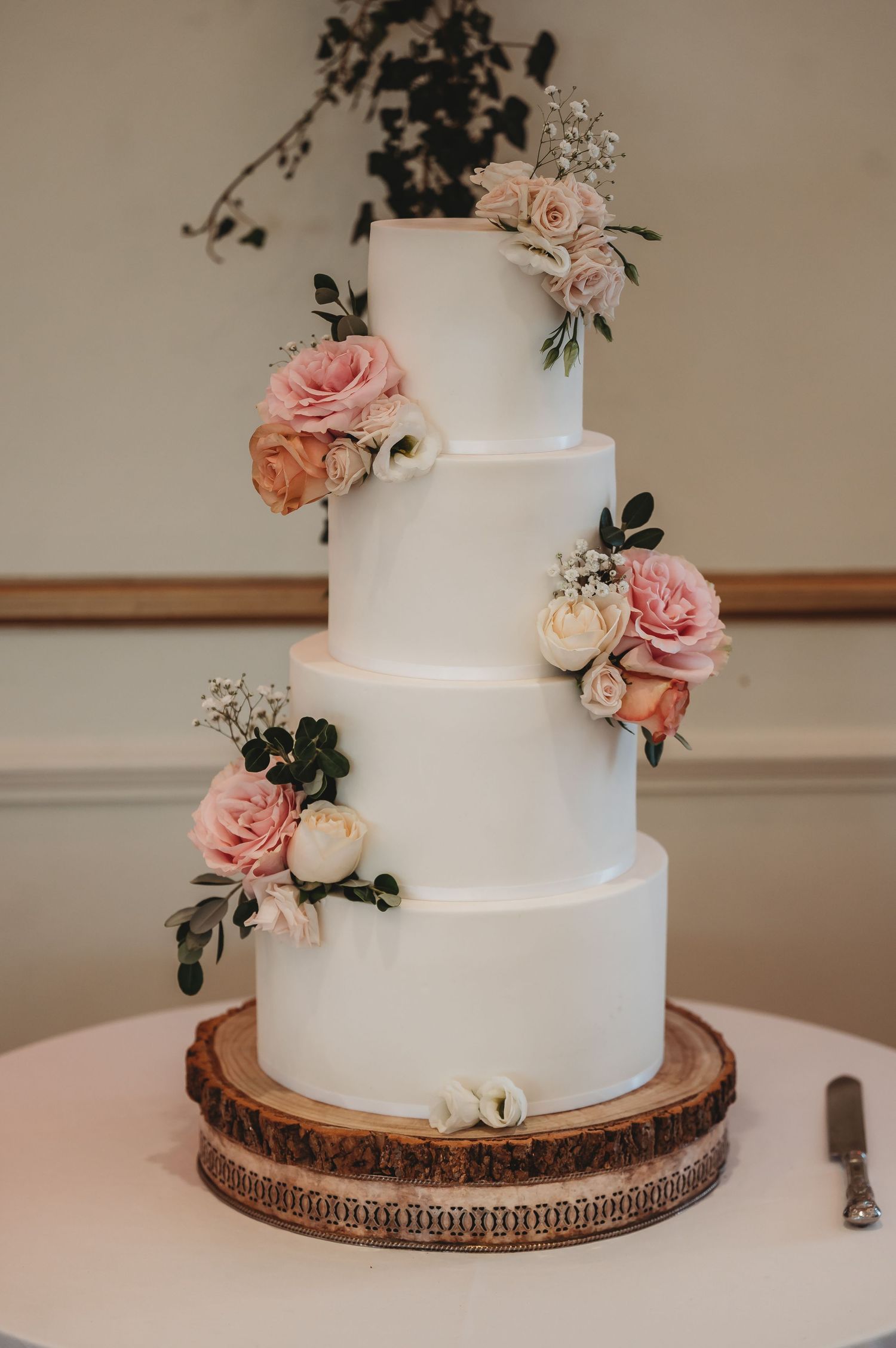 Elegant four-tier white wedding cake decorated with pink and peach roses on a rustic wooden cake stand.