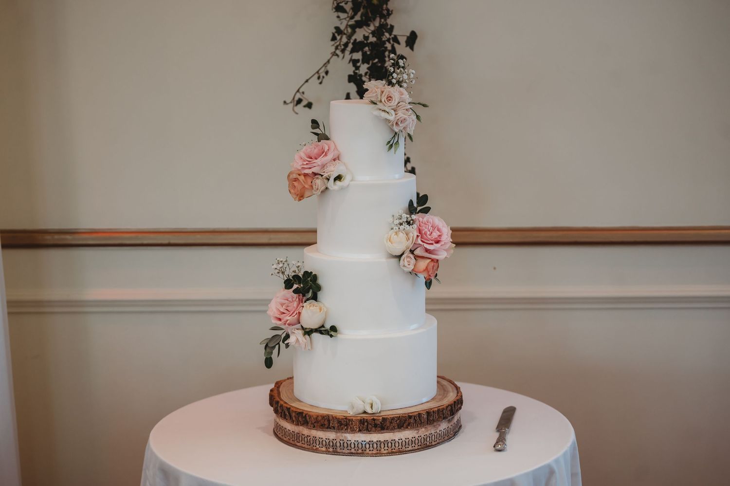 Elegant three-tier white wedding cake decorated with pink roses and greenery on wooden cake stand.