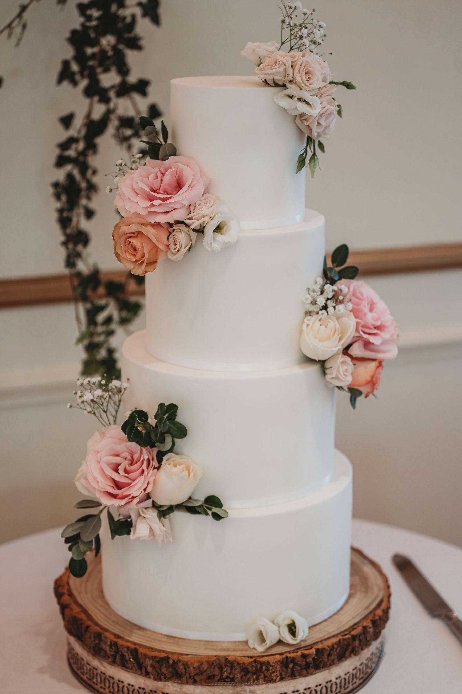 Classic white tiered wedding cake adorned with blush pink roses and greenery on a wood slice base.