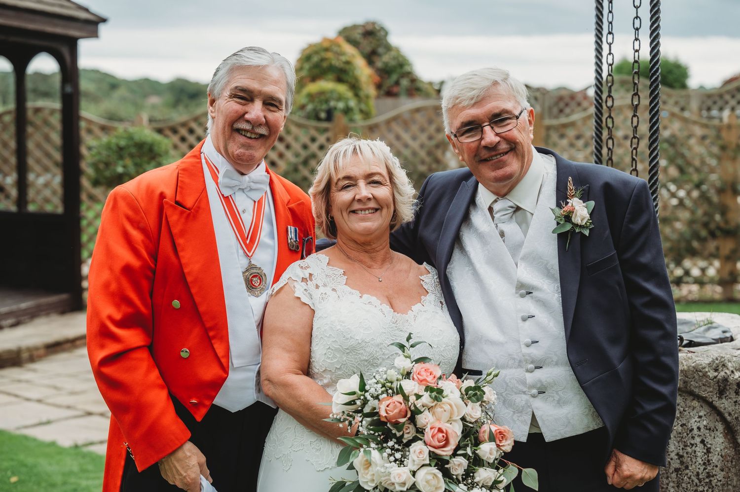 Three people in formal attire pose together outdoors with the person in the middle holding a pink floral bouquet.