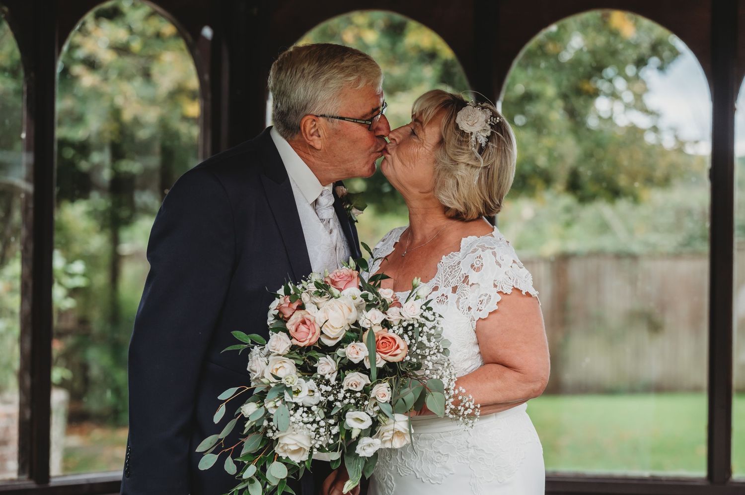 A romantic wedding couple shares a tender kiss beneath arched architecture while holding a pink and white floral bouquet.