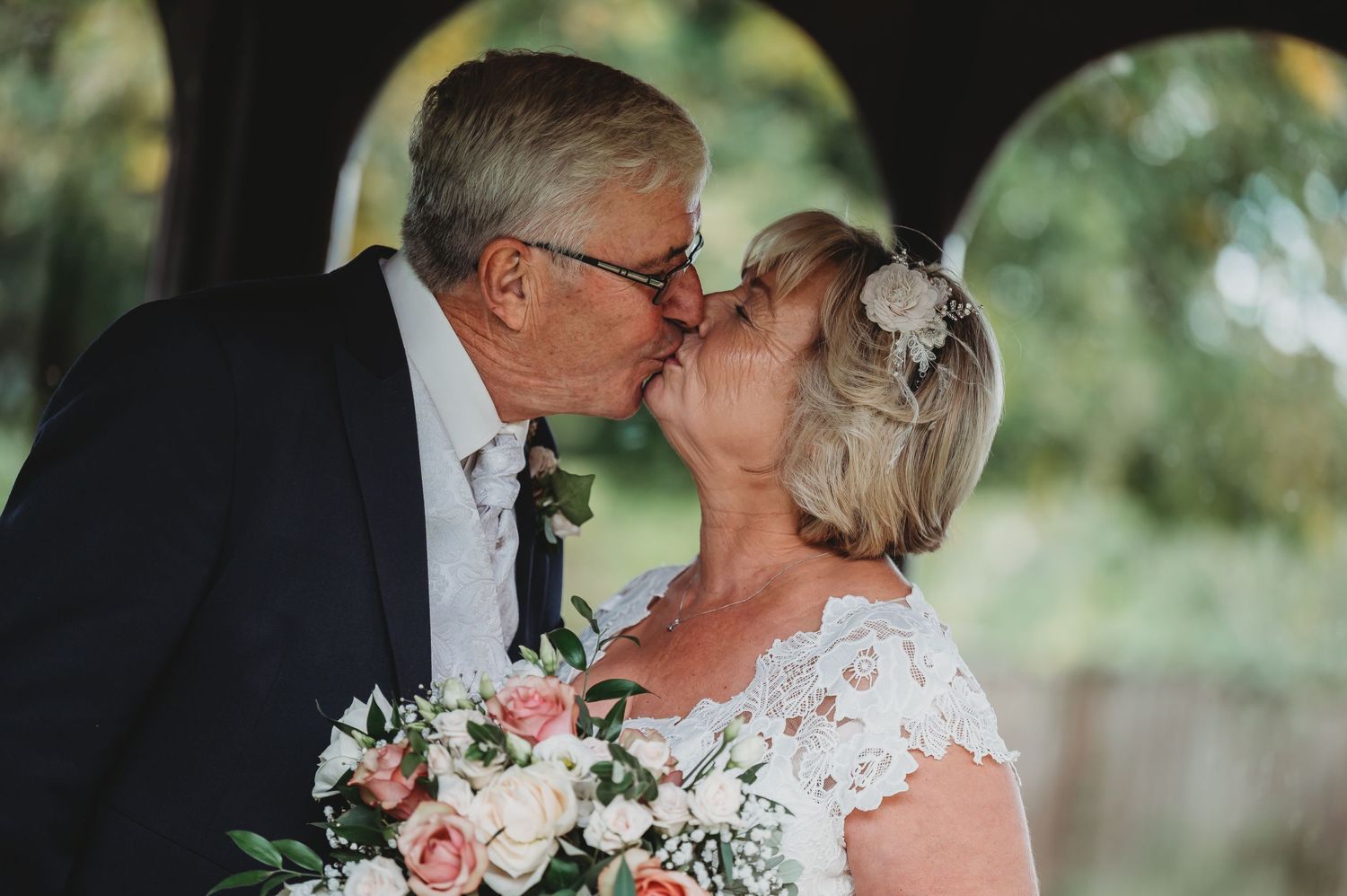 A romantic wedding couple shares a tender kiss while holding a pink and white floral bouquet in an outdoor garden setting.