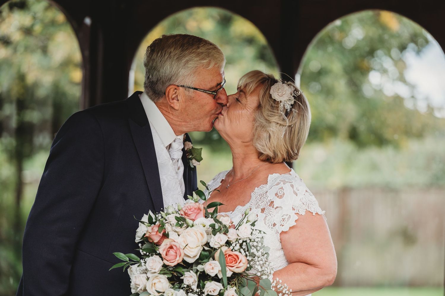 A romantic wedding kiss moment captured outdoors with a pink and white bridal bouquet.