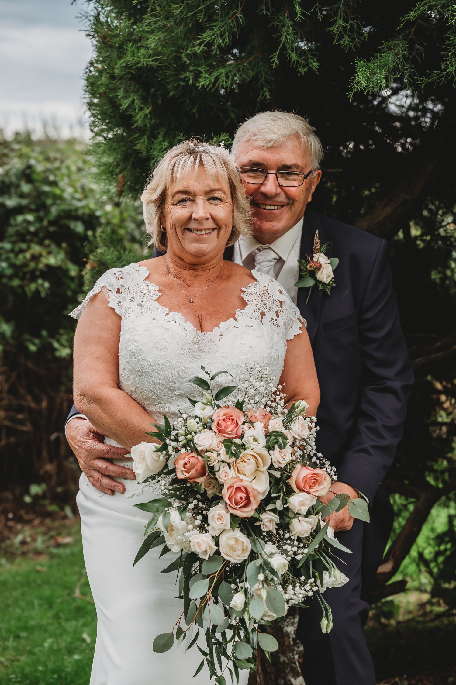 Happy couple in wedding attire smile together outdoors with a beautiful pink and white floral bouquet.