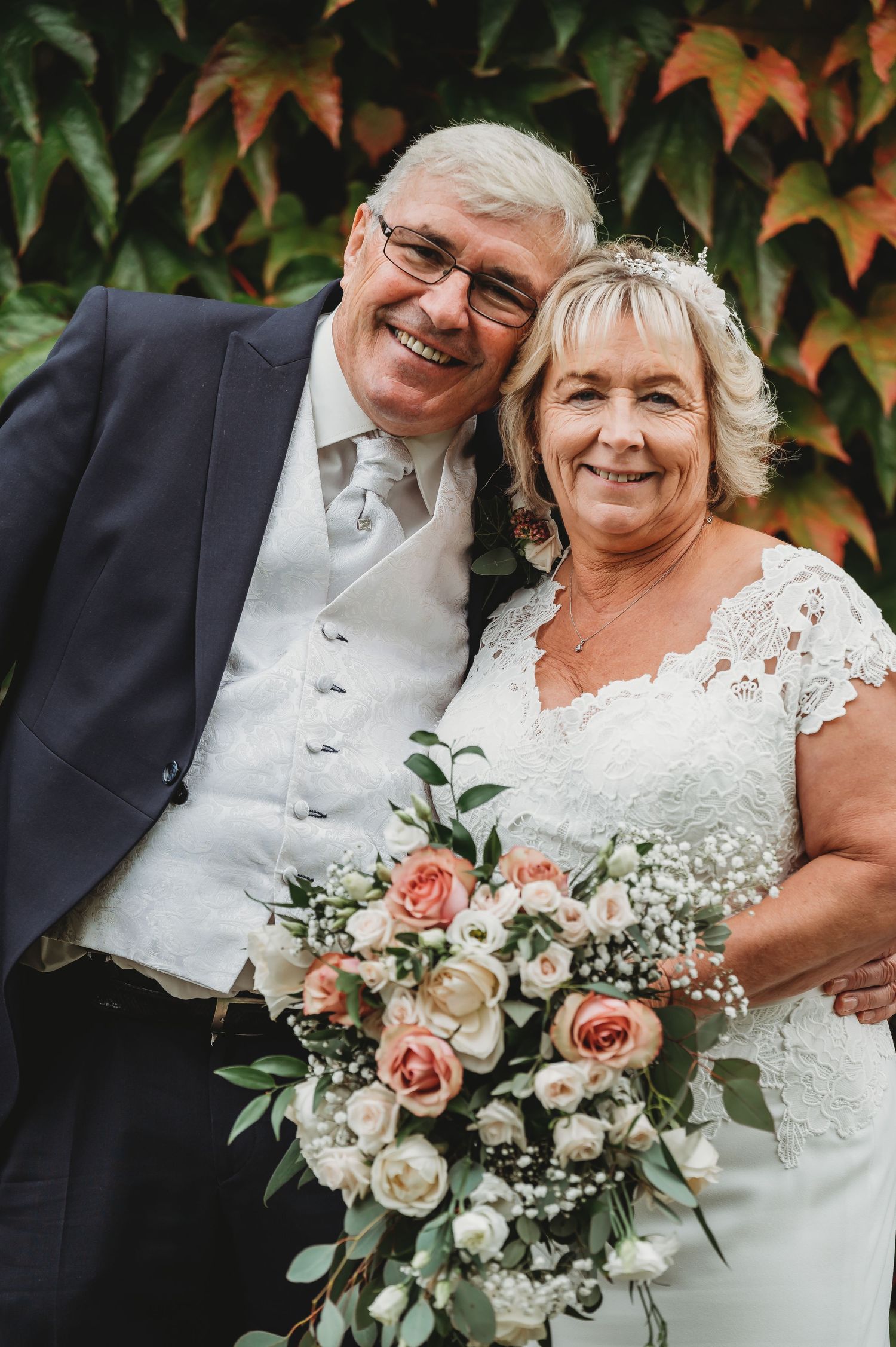 A joyful couple in wedding attire pose outdoors with a beautiful bouquet of pink roses and white flowers against autumn foliage.