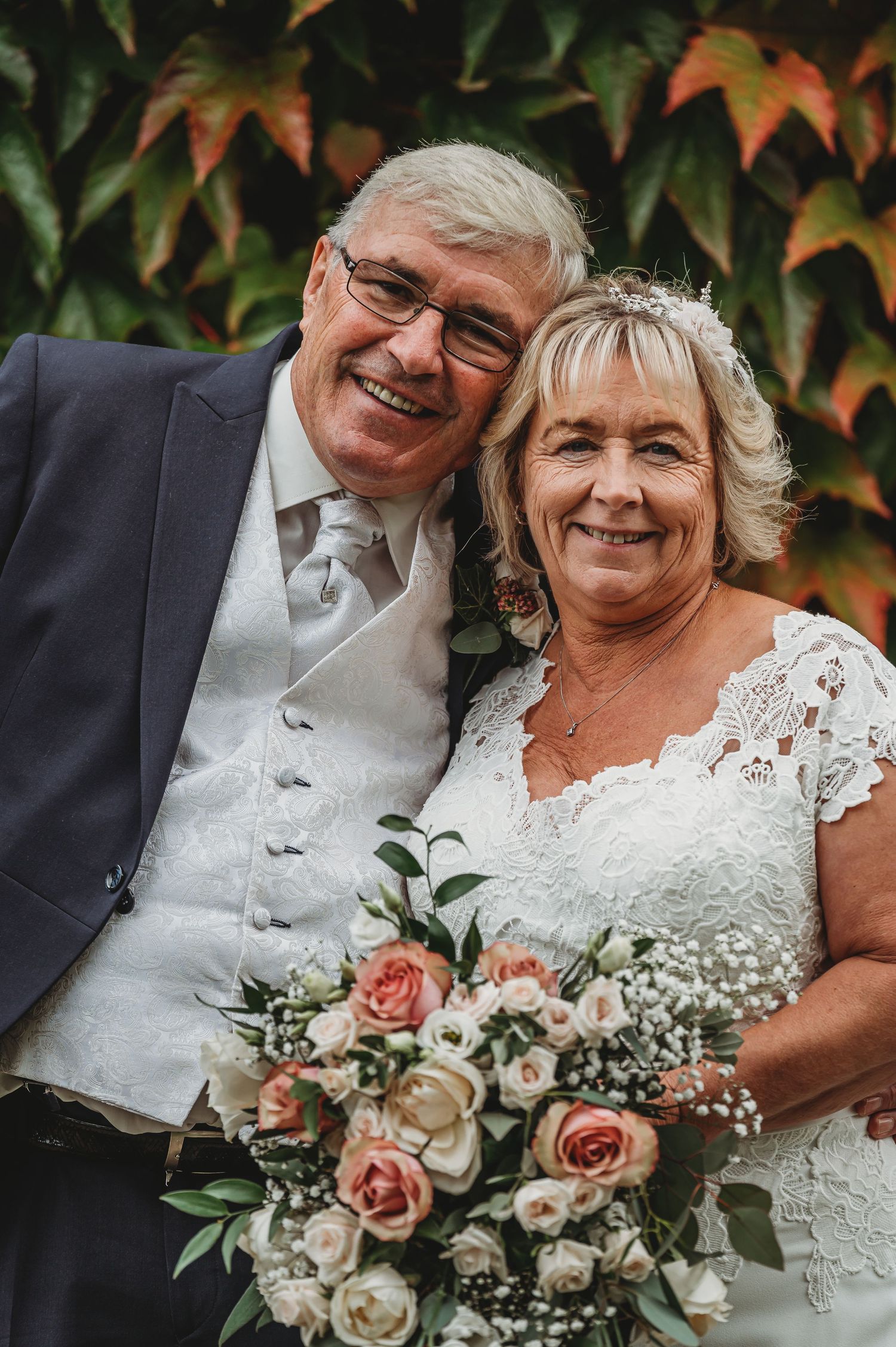 Happy couple in formal attire smiling outdoors with autumn leaves and pink roses in bridal bouquet.