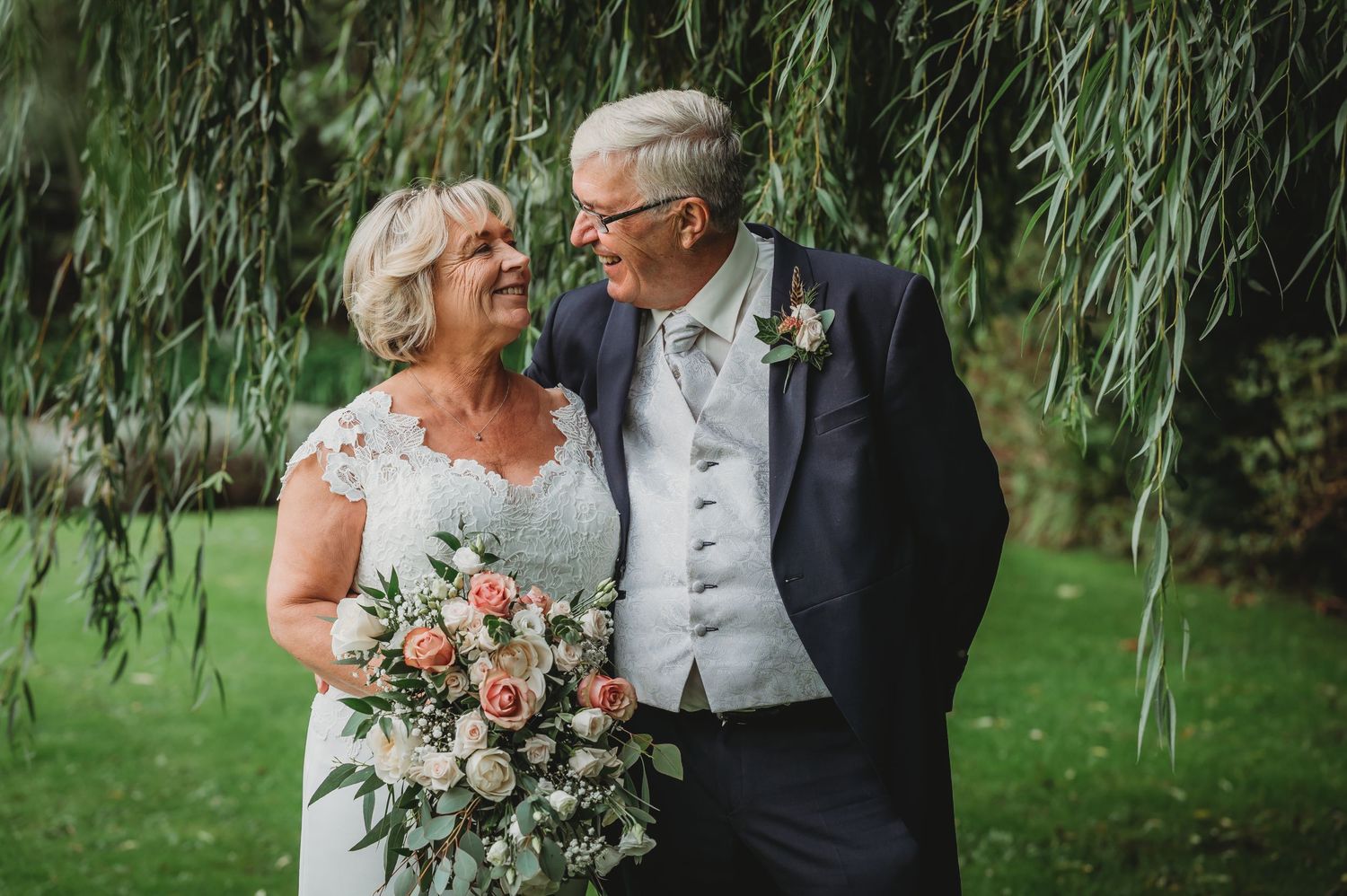 A happy elderly couple shares a tender moment under weeping willow branches during their wedding celebration.