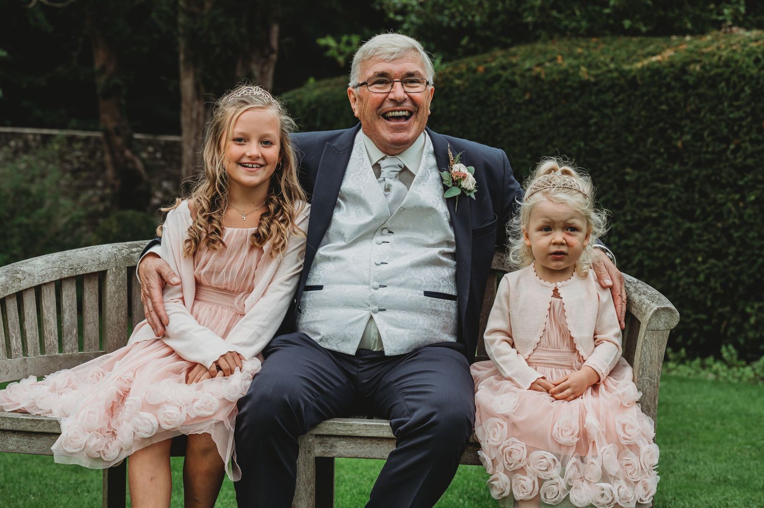 Adults and children in formal attire smile while sitting together on a wooden bench in a garden setting.