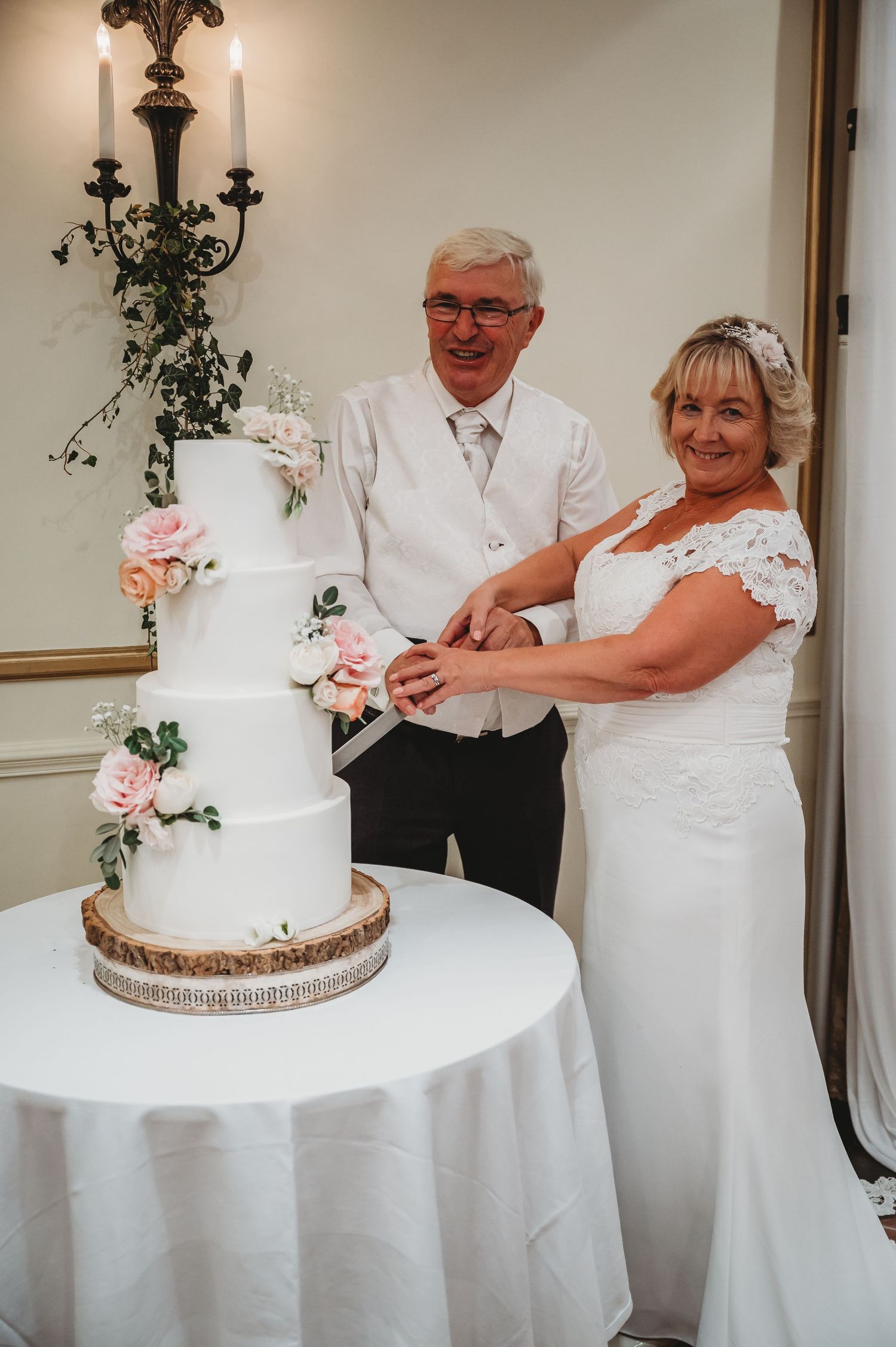 A couple in elegant white attire cuts their four-tier wedding cake decorated with pink roses on a round table.