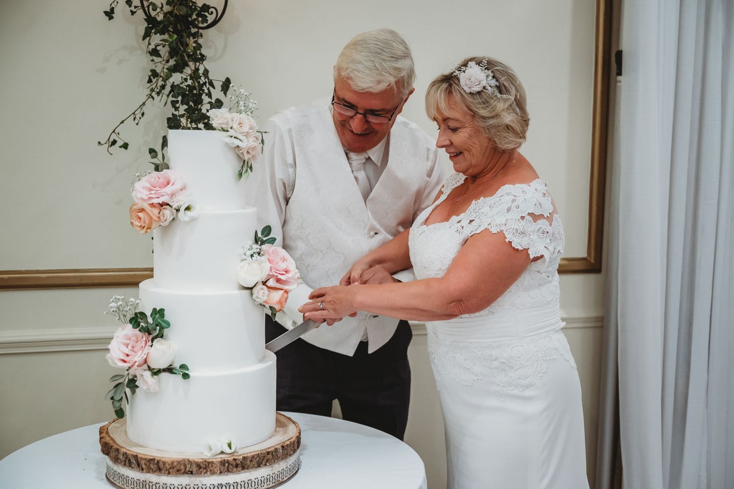 An elegant couple cut their white wedding cake decorated with pink roses and greenery at their celebration.