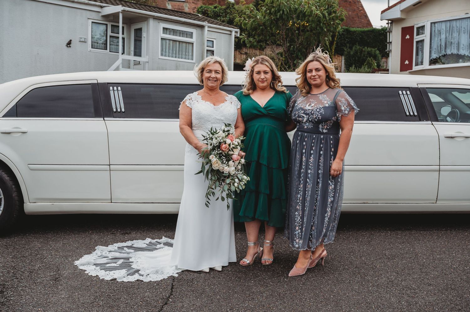 Three people stand smiling in front of a white stretch limousine at a special event.