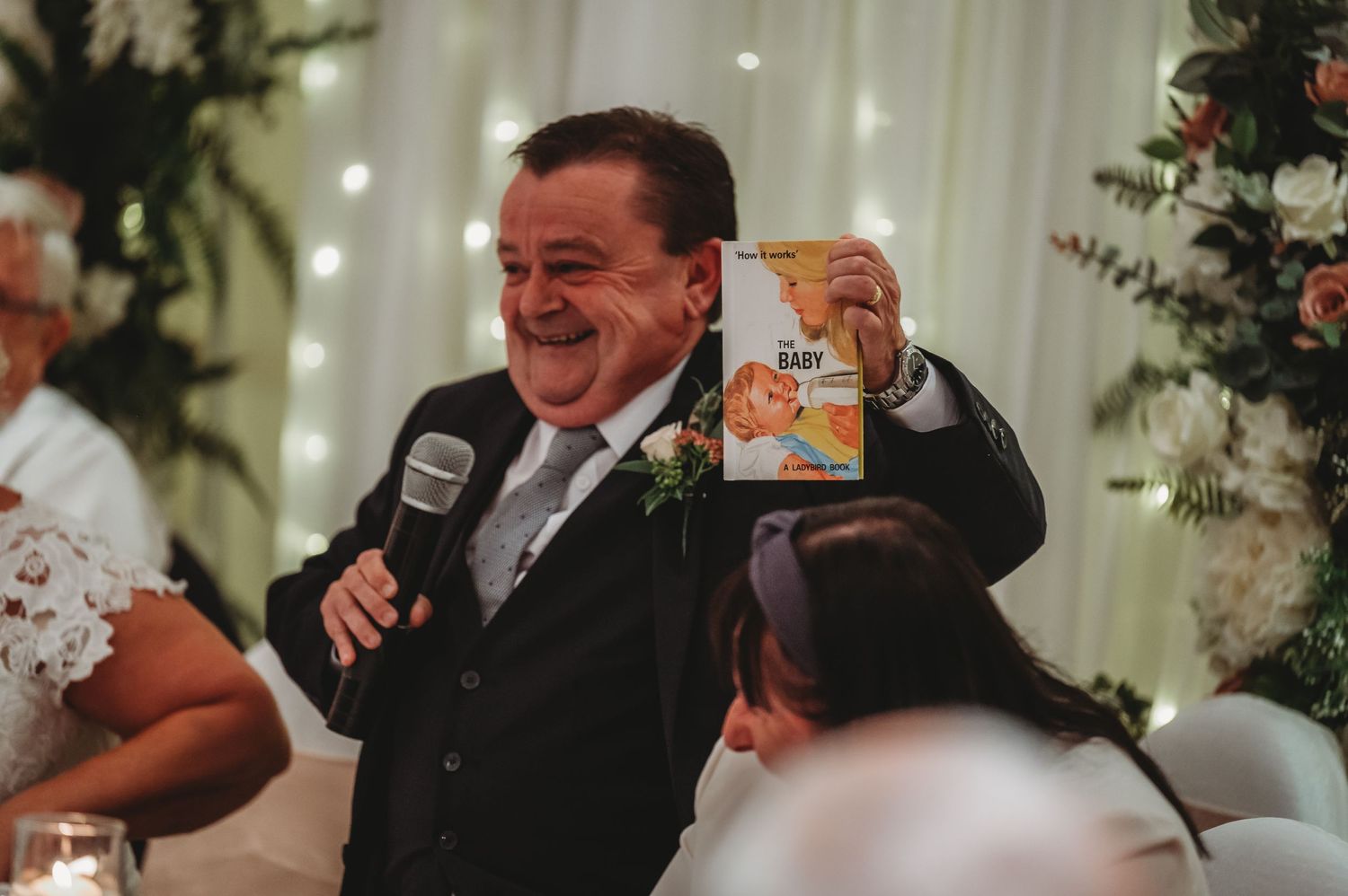 Someone in a black suit makes a speech while holding up a Golden Grahams cereal box at a reception event.