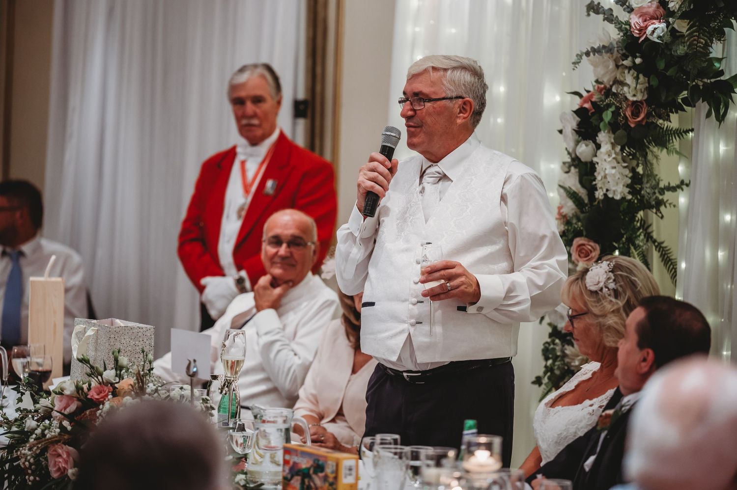 A speaker addresses guests at an elegant wedding reception with holiday decorations and white table settings.