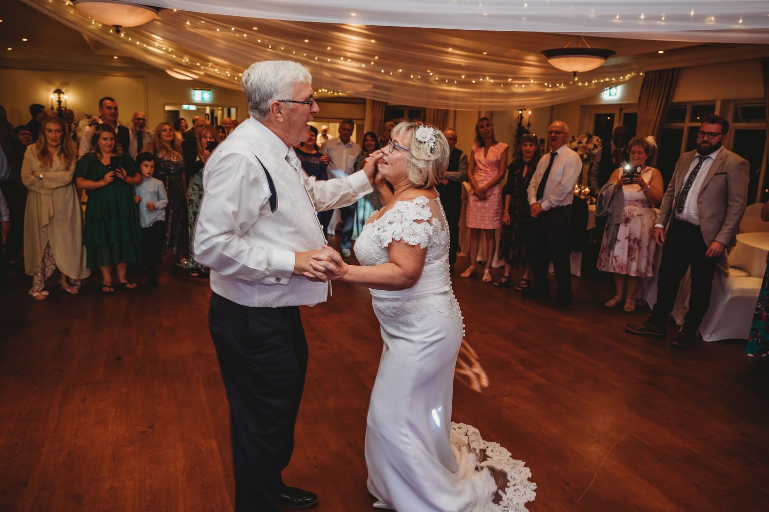 People dancing at an elegant wedding reception on a polished wood dance floor under warm lighting.