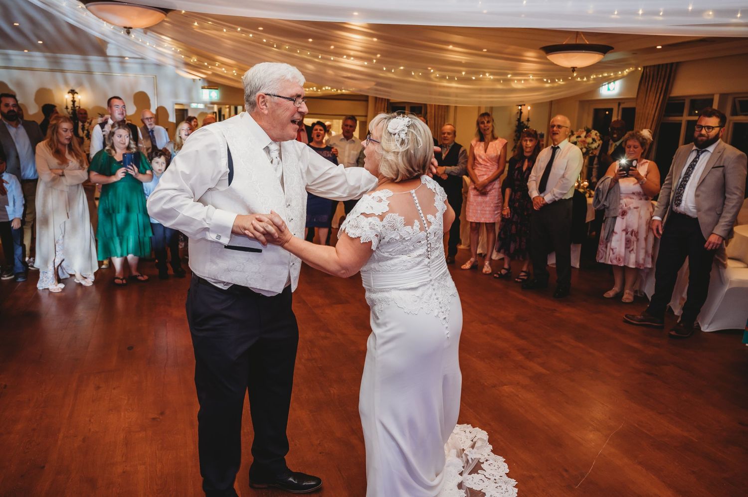 Two people enjoy a special dance together on a polished wooden dance floor while wedding guests watch from the edges.