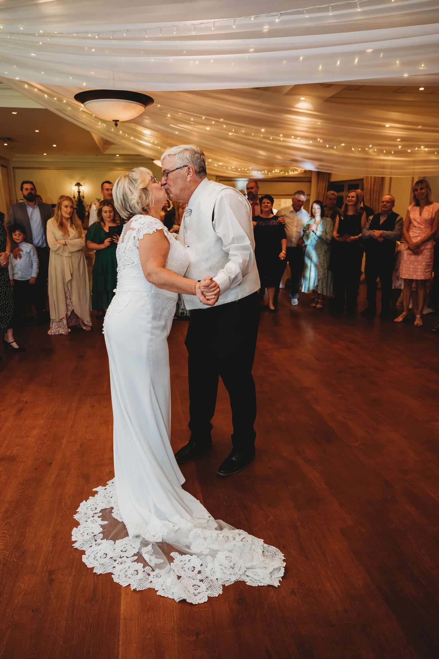 A couple shares a tender dance moment under twinkling lights on a polished wooden dance floor at their wedding reception.
