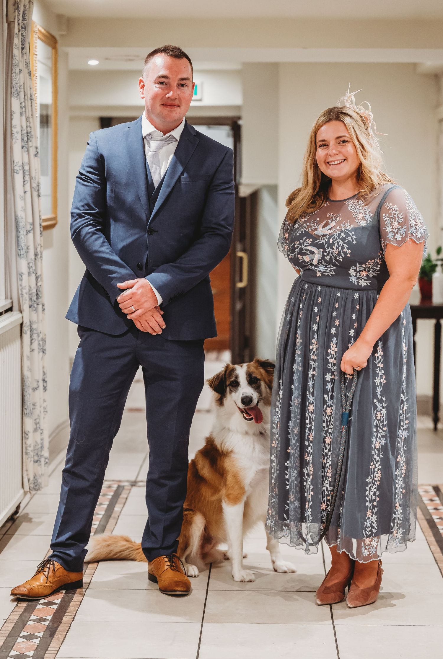 A couple in formal attire stands in a hallway with their Australian Shepherd dog between them.