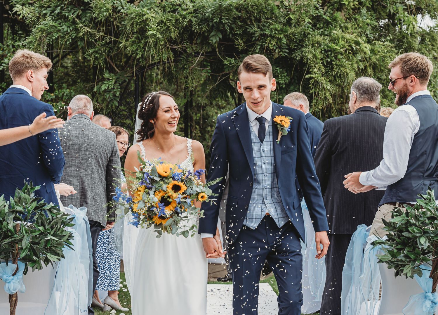 Wedding guests celebrating by throwing confetti over a couple holding bright sunflower bouquet in an outdoor garden setting.