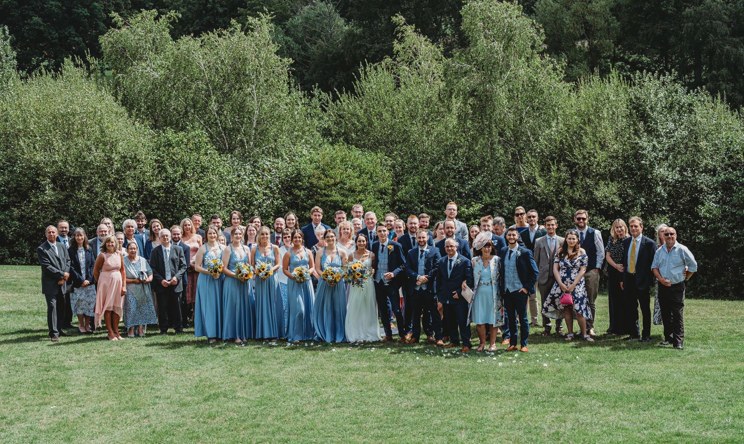 Wedding party in coordinated blue and gray attire poses together on lush green lawn for formal group photo.