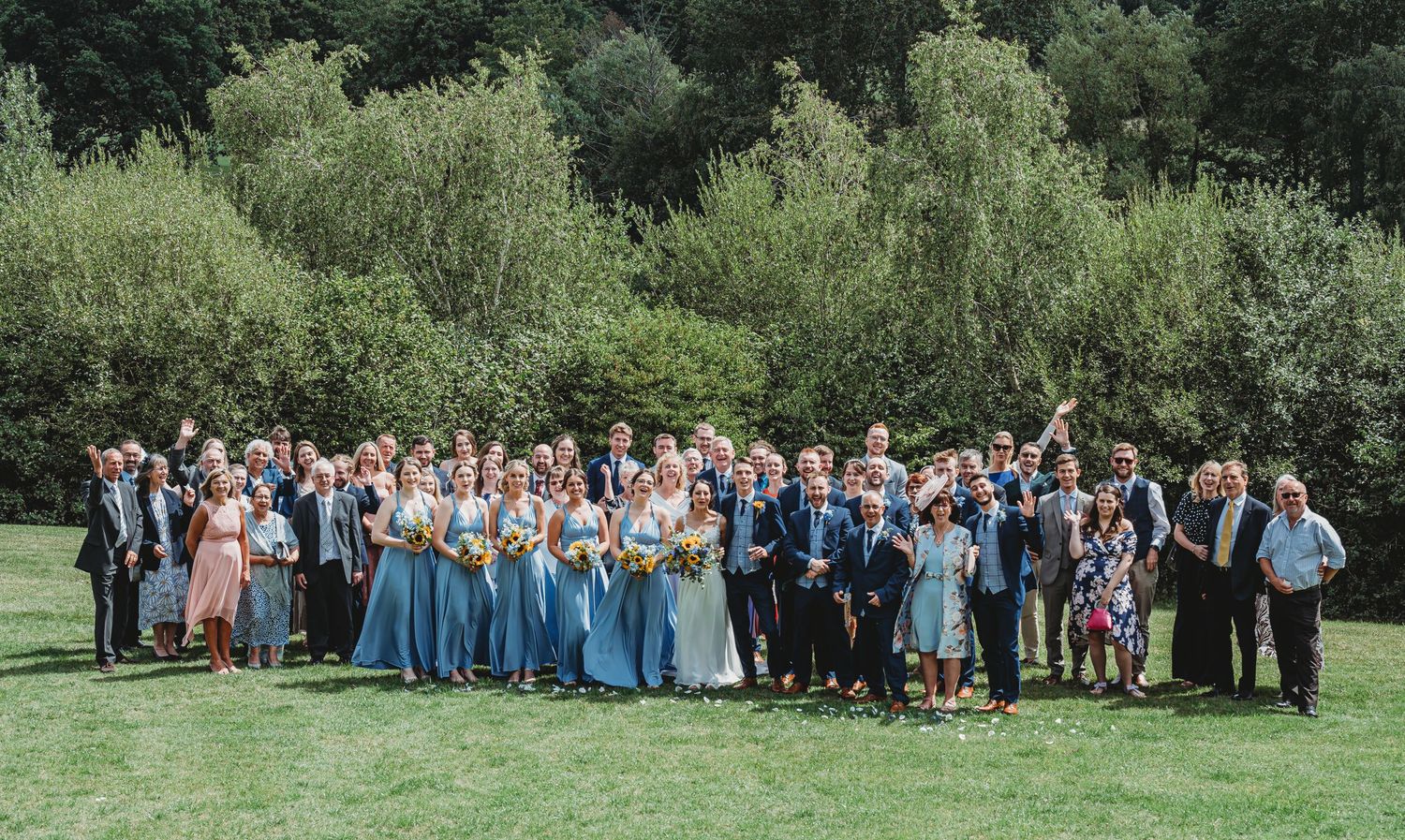 Large wedding party poses together outdoors on green lawn with lush trees in background.