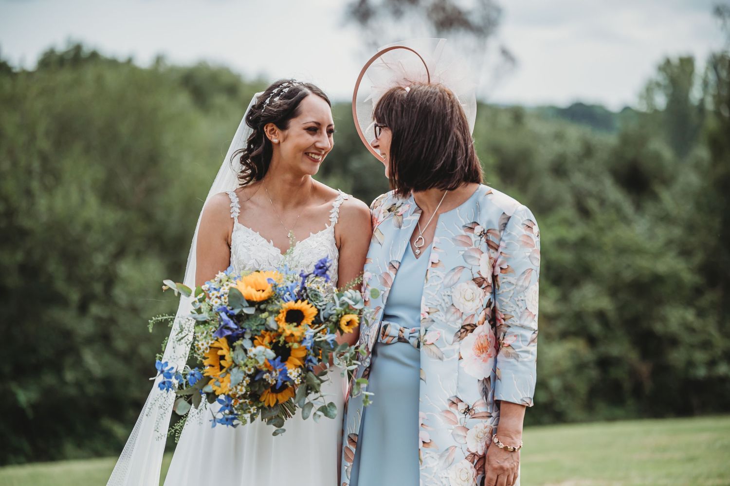 Two wedding guests share a joyful moment outdoors, with one holding a vibrant sunflower and blue floral bouquet.
