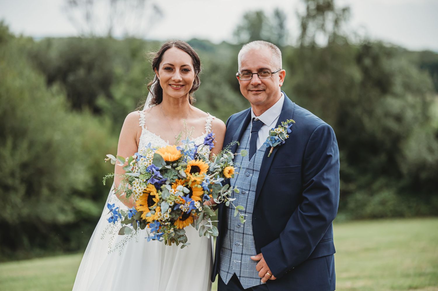 Two people pose together at an outdoor wedding with a colorful sunflower bouquet against a green landscape.
