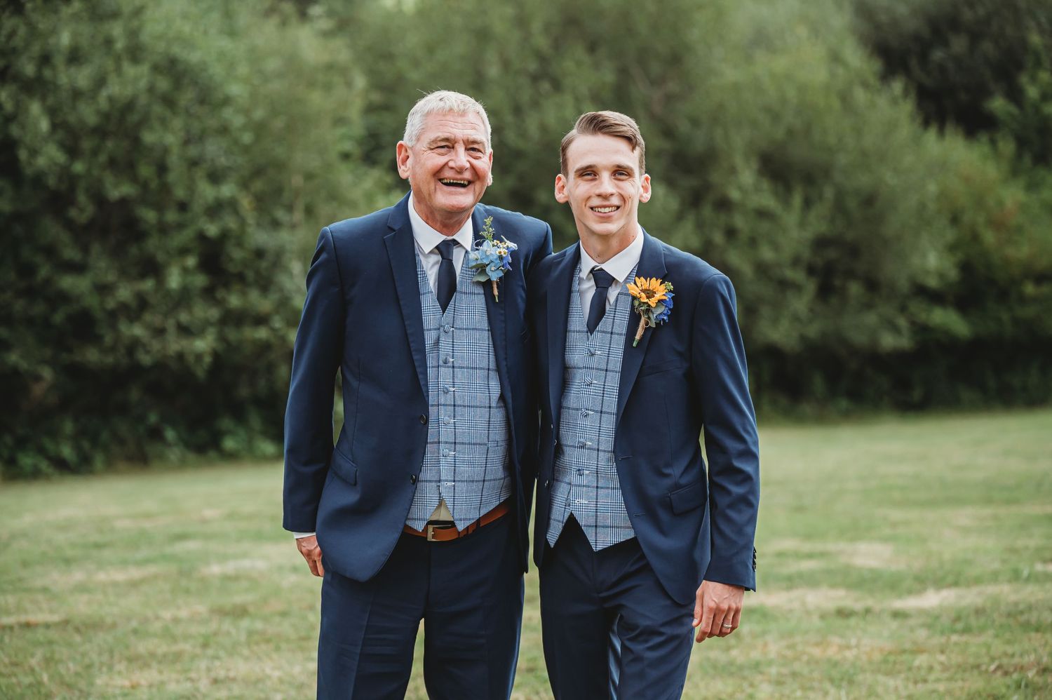 Two men in matching navy suits and checkered waistcoats smile together during an outdoor wedding celebration.