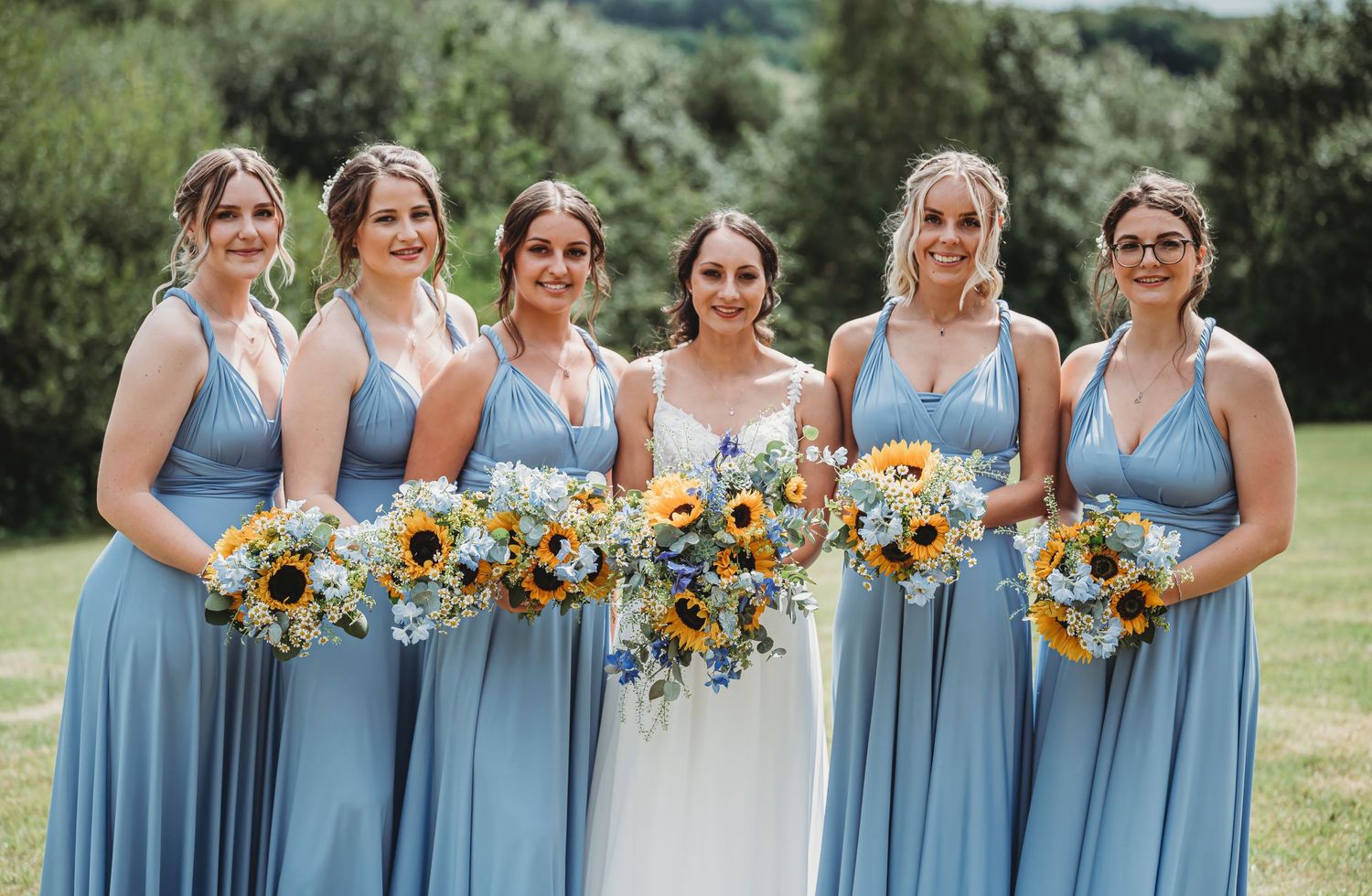 A wedding party wearing light blue bridesmaid dresses poses outdoors holding vibrant sunflower bouquets.