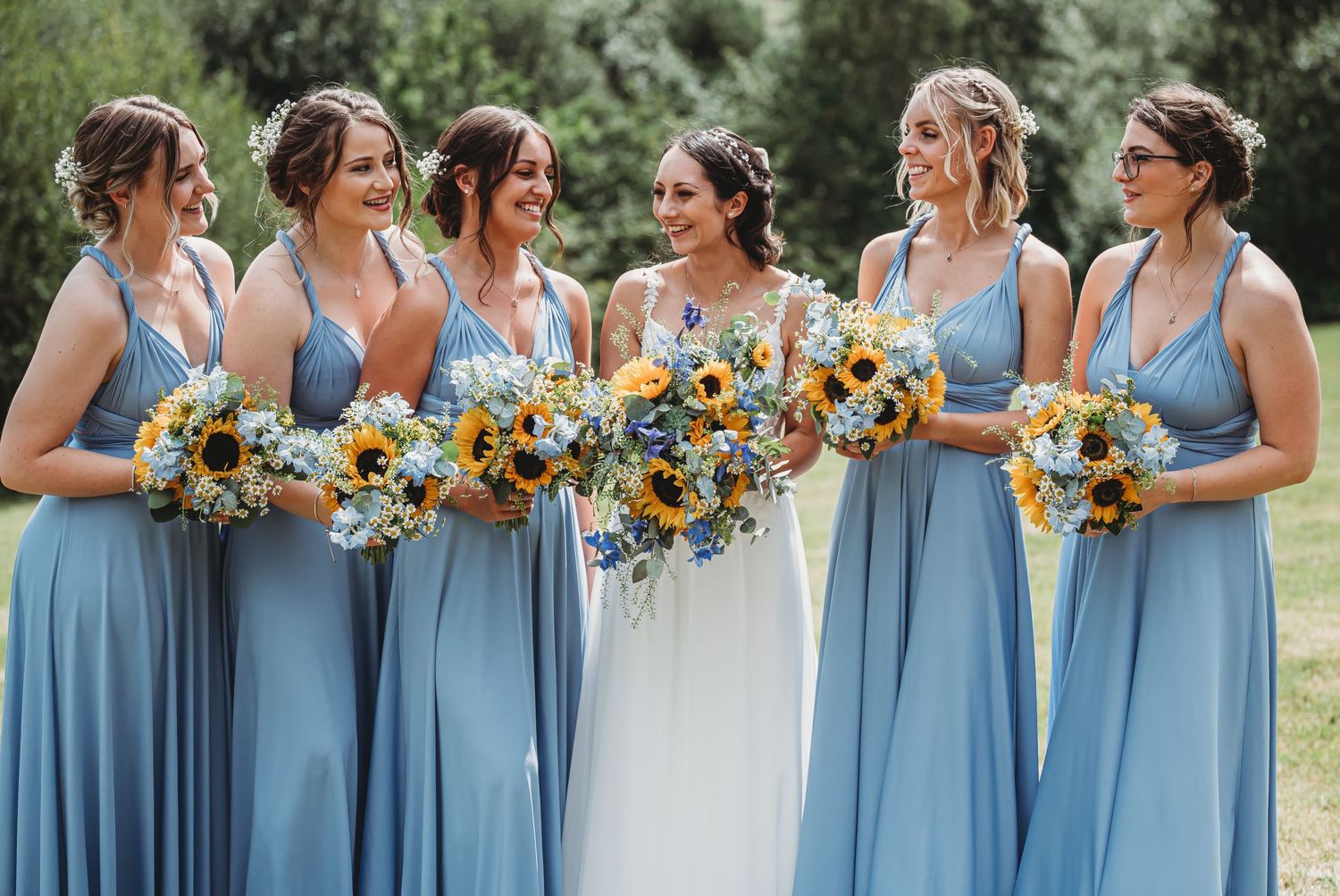 Wedding party wearing dusty blue bridesmaid dresses holding vibrant yellow sunflower bouquets in outdoor garden setting.
