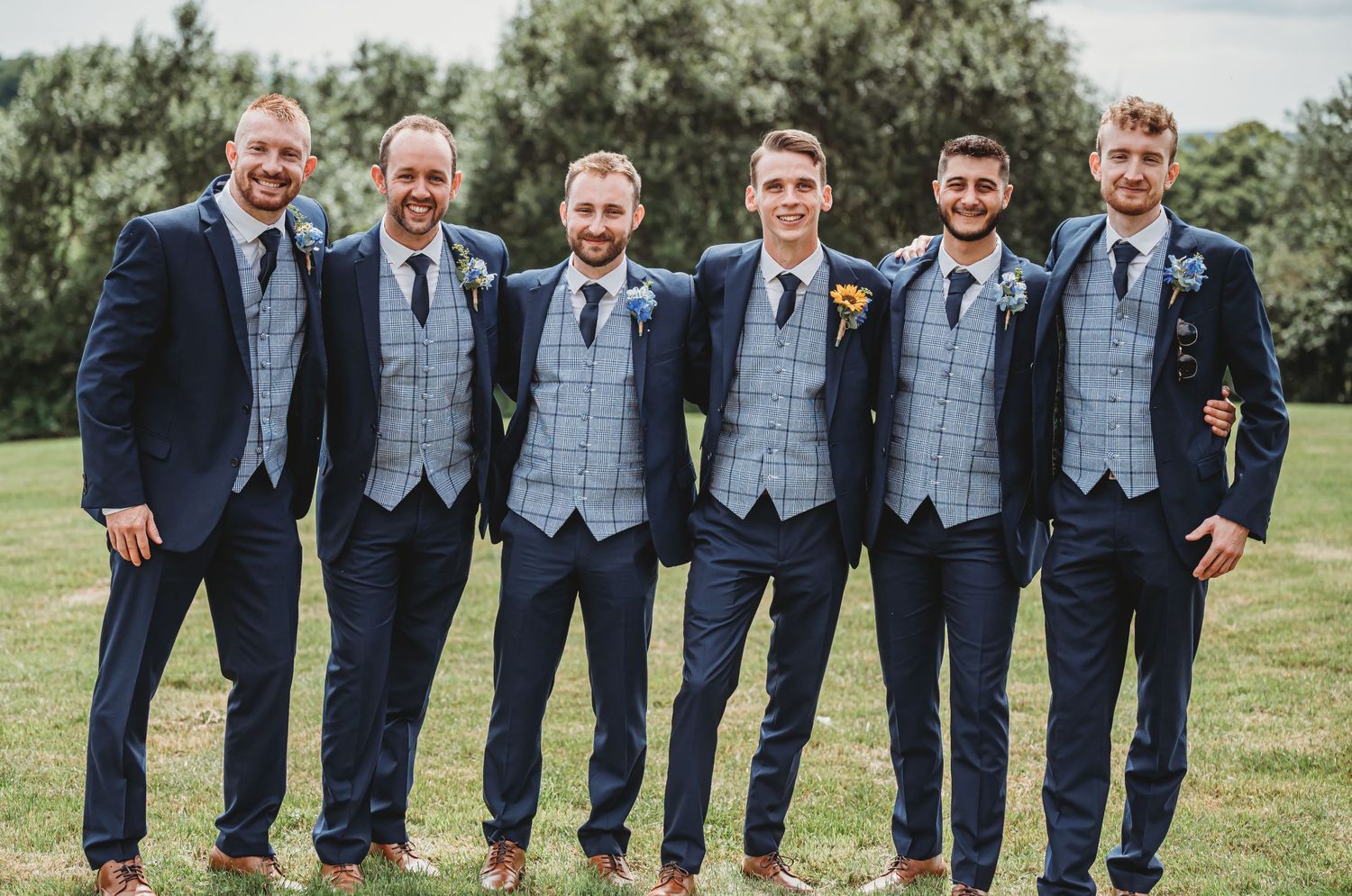 Groomsmen wearing coordinated navy suits with light plaid vests pose together on green lawn at outdoor wedding.