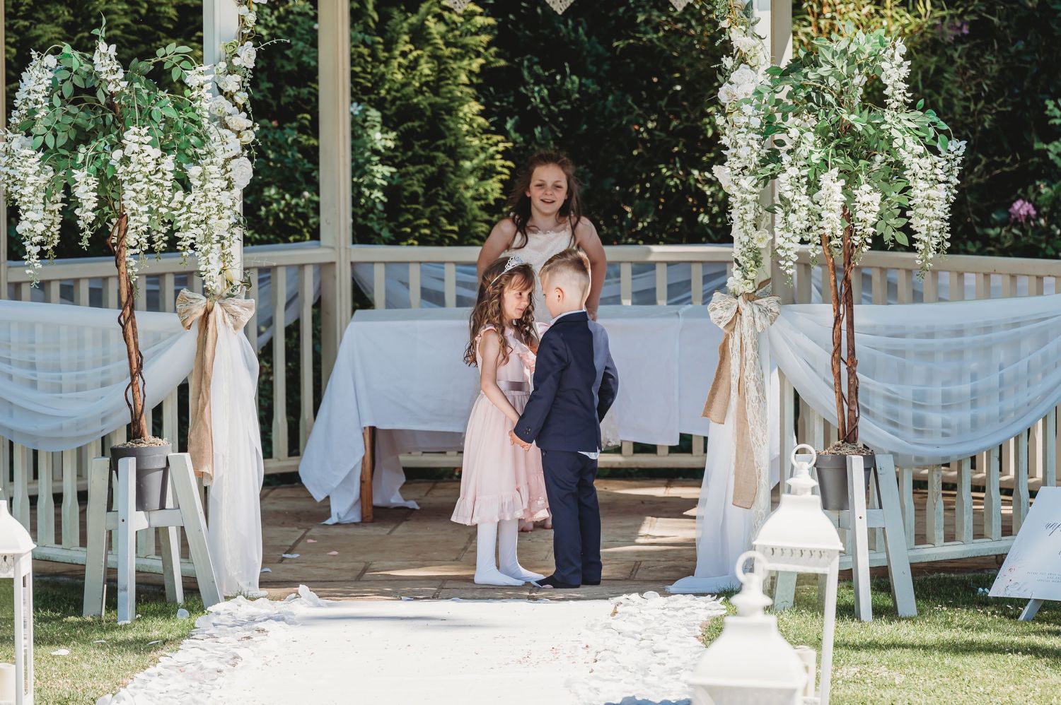 A rustic outdoor wedding ceremony setup with white floral arrangements and draped fabric decorating a wooden gazebo.