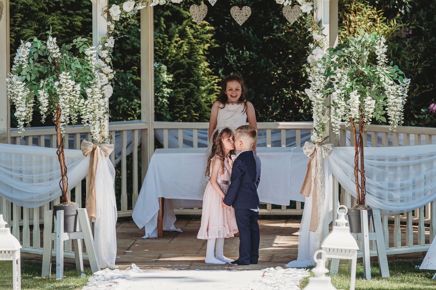Elegant outdoor wedding setup with white decor, flowing fabric draping chairs, and decorative trees at a garden ceremony location.
